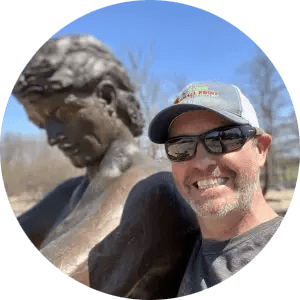Photo of Chris standing in front of a statue while wearing a baseball cap and sunglasses.