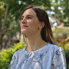 Woman standing in front of trees and shrubs wearing a white floral shirt.