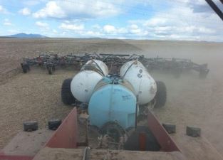 View from inside a vehicle showing a tractor with large tanks and plowing equipment working in a dry, dusty field under a cloudy sky.