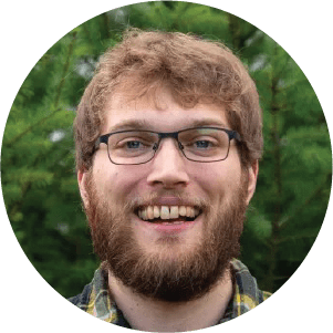 A smiling young man with a beard and glasses, wearing a plaid shirt, standing in front of a green, forested background.