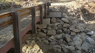 Wooden bridge under construction with stacked rocks and logs on uneven ground.