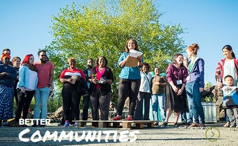 A diverse group of people listens to a woman speaking outdoors, with the text "better communities" overlaid on the image.