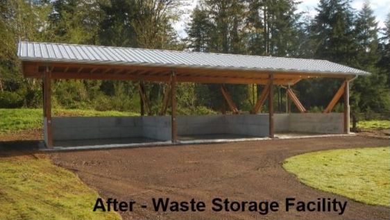 Outdoor waste storage facility with a metal roof supported by wooden beams and concrete walls, situated in a grassy area with trees in the background.