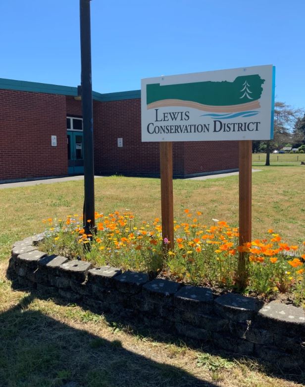 A sign for lewis conservation district stands in a grassy area with orange wildflowers, in front of a brick building under a clear blue sky.