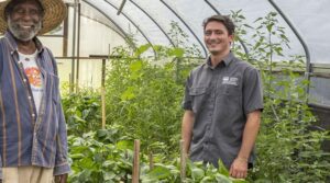 Two men, one elderly with a hat and another younger in a work shirt, smiling inside a greenhouse surrounded by plants.