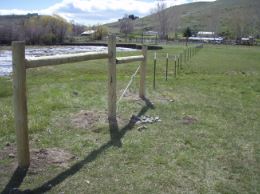 A wooden fence extends into a green field near a flowing river, with a hill and buildings in the background under a cloudy sky.