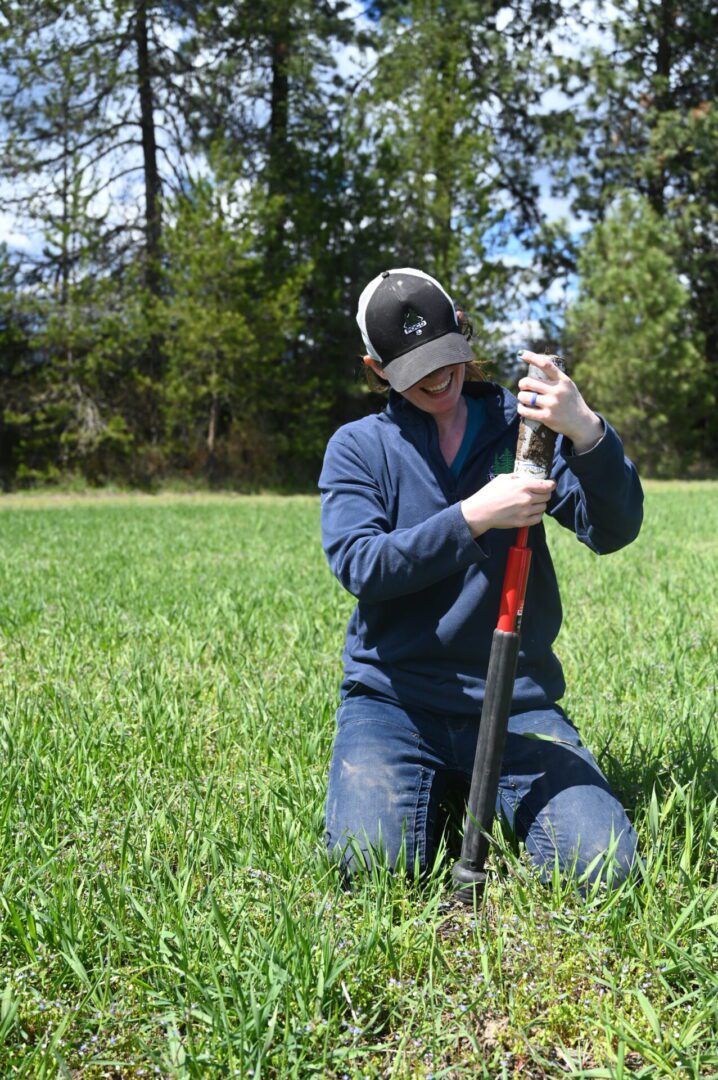 A person in a baseball cap and sunglasses using a soil sampling tool in a lush green field with trees in the background.