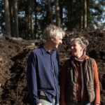 Hilary-McMullen_Midnights-Farm_97A1439 An older couple smiling at each other, standing by a large pile of mulch in a wooded area on a sunny day.