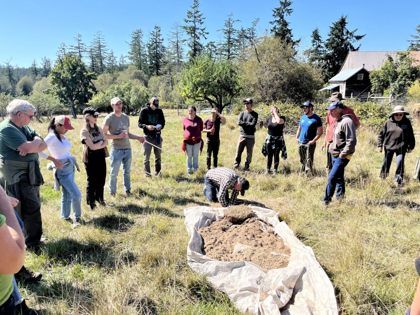 A group of adults observing a demonstration in a grassy field, with one person kneeling by a pile of soil on a tarp, beneath a clear blue sky.