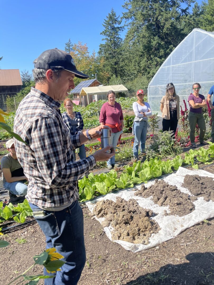 IMG_4826 A man demonstrating gardening techniques to a group in a community garden, with a greenhouse in the background.