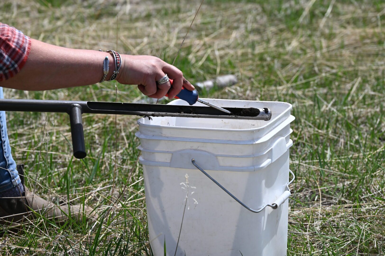 A person in a plaid shirt scooping soil with a trowel into a white bucket in a grassy field.