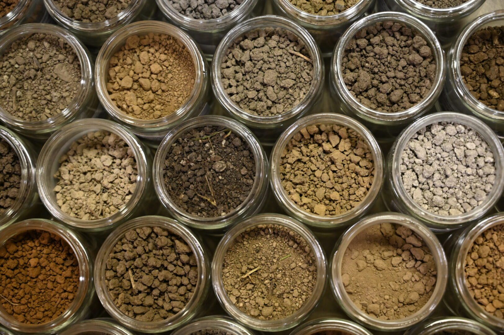 Assorted granular materials in shades of gray and brown, displayed in transparent cylindrical containers arranged in rows.