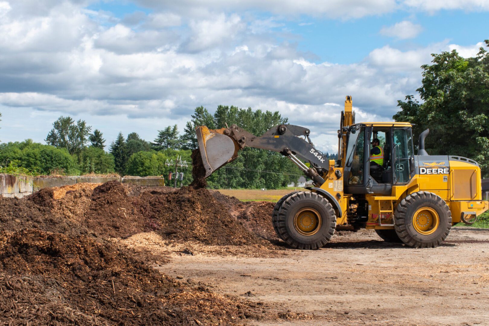 A yellow bulldozer moves a scoop of compost, surrounded by green trees under a partly cloudy sky. Worker visible in the cab.
