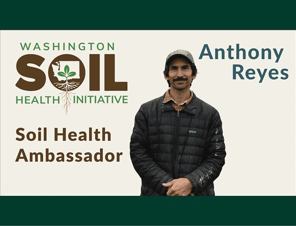 A promotional graphic for the Washington Soil Health Initiative featuring the program’s logo with a plant and roots inside the word “SOIL.” The text reads “Soil Health Ambassador” on the left and “Anthony Reyes” on the right. In the center, a person wearing a dark quilted jacket, plaid shirt, and green cap stands against a light background.