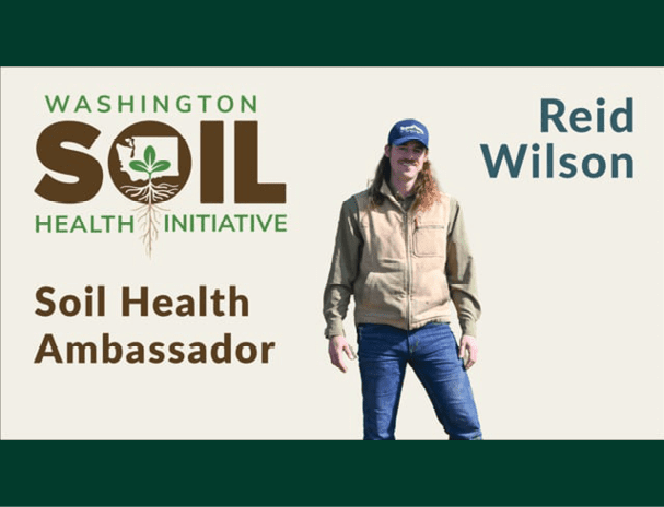 A promotional graphic for the Washington Soil Health Initiative featuring its logo with a plant and roots inside the word “SOIL.” The text on the left reads “Soil Health Ambassador,” and on the right, “Reid Wilson.” In the center, a person is standing wearing a tan vest over a long-sleeve shirt, blue jeans, and a dark baseball cap. The background is light with dark green borders at the top and bottom.