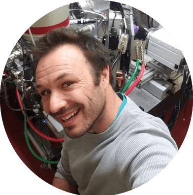 A person wearing a light gray long-sleeve shirt is seated in front of complex laboratory equipment. The setup includes metallic components, wires, and tubing in red, green, and black colors, suggesting a scientific or technical environment such as an analytical instrument or experimental apparatus. The background shows multiple connected devices and machinery with a mix of shiny metal surfaces and control panels.