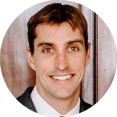 A closeup image of Gabe wearing a white shirt and a black jacket, standing in front of a wood paneled wall.