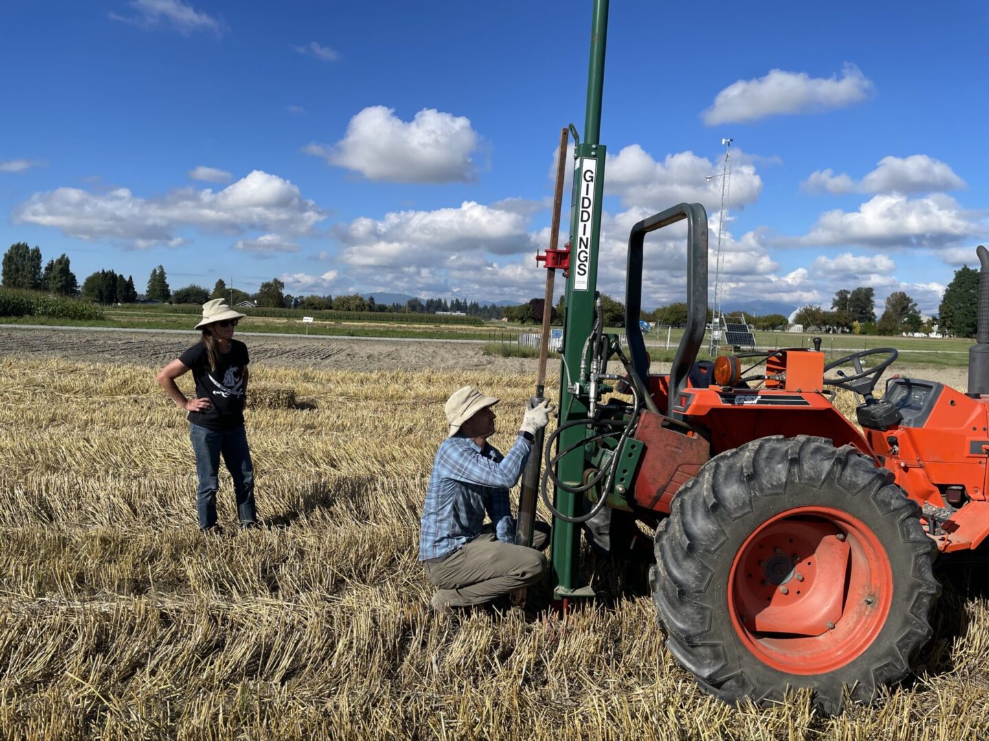 Two people are working in a harvested field under a bright blue sky with scattered clouds. One person is standing and observing while the other kneels next to a large orange tractor equipped with a green soil sampling probe labeled “GIDDINGS.” The tractor has a large rear tire and is positioned on dry, cut crop residue. In the background, there are rows of crops, trees, and distant mountains. The scene suggests soil sampling or field research activity.