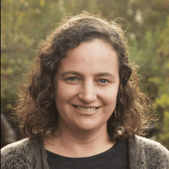 A woman standing in front of a row of trees in a black shirt and grey cardigan.