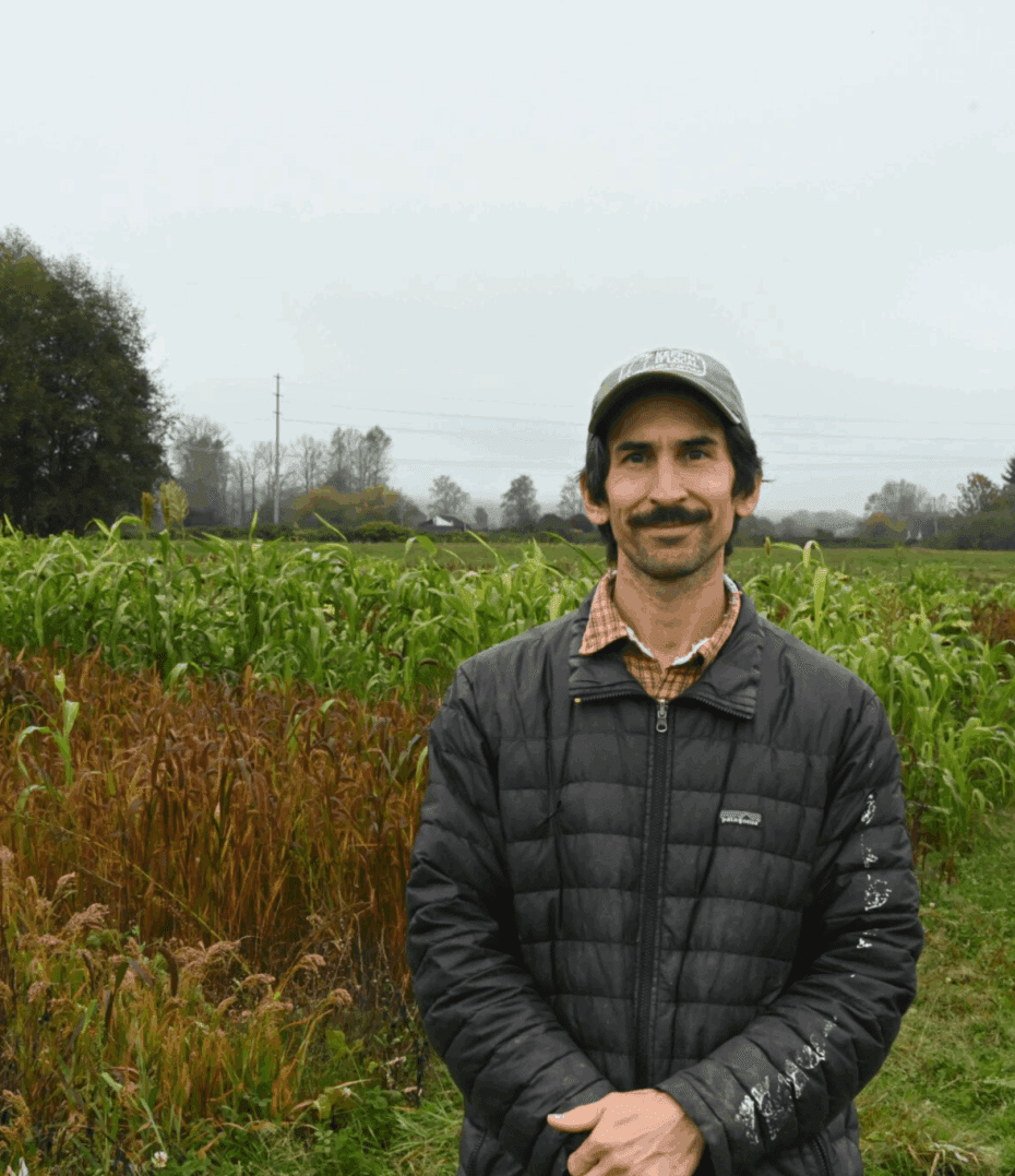 Anthony Reyes wears a black jacket and a baseball cap standing in front of a field at Oxbow Farm under an overcast sky.