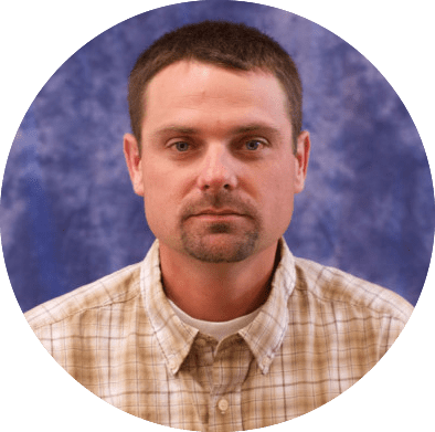 A circular portrait photo showing a person wearing a light brown plaid button-up shirt over a white undershirt. The background is a mottled blue and purple studio backdrop. The image has a professional headshot style.
