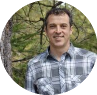 A person wearing a gray plaid button-up shirt stands outdoors in front of a tree trunk and green foliage. The background shows a natural setting with branches and leaves, suggesting a forest or wooded area. The individual is positioned centrally in the frame, and the image is cropped in a circular format.