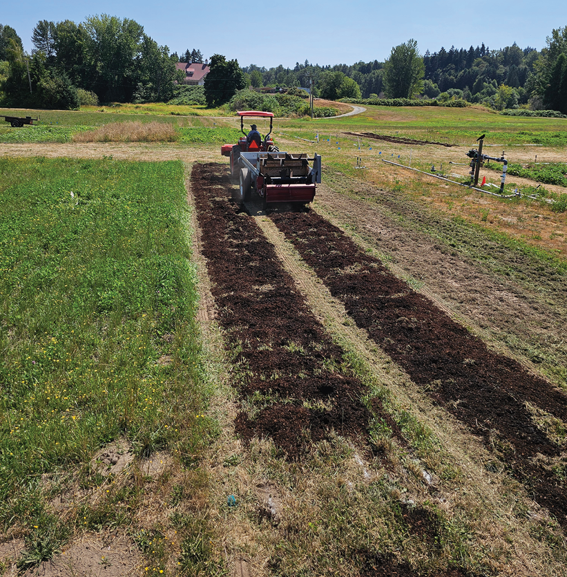 A tractor with a rear-mounted spreader moves along two prepared rows in a grassy field, applying a dark organic material, likely compost or mulch, onto the soil. The rows of applied material contrast with the surrounding green grass and lighter bare soil. In the background, there are trees, a winding dirt path, and a small building partially visible among the foliage under a clear blue sky, suggesting a farm or research site in summer.