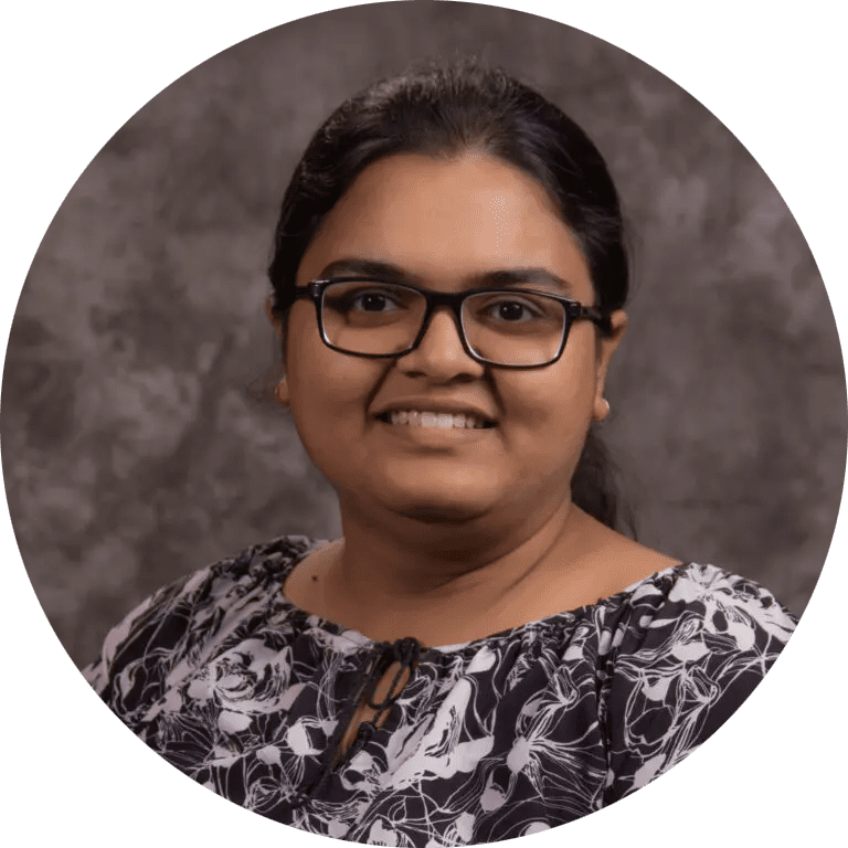 A person is shown in a professional headshot against a textured gray backdrop. The individual is wearing a black blouse with a white floral pattern and a small keyhole neckline detail. The hair is pulled back, and the photo is cropped in a circular format, focusing on the upper body and shoulders.