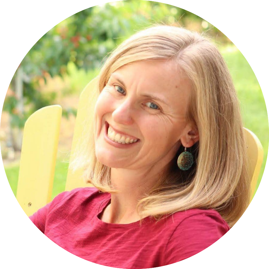 A person with shoulder-length blonde hair is seated outdoors on a yellow chair. The individual is wearing a maroon shirt and dark earrings. The background shows green foliage and blurred plants, suggesting a garden or natural setting on a sunny day.