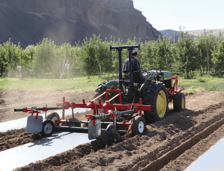 A tractor pulls a red implement designed for laying plastic mulch over raised soil beds in an orchard. The machine has multiple wheels and metal arms guiding the plastic sheets onto the soil, creating smooth, covered rows. The tractor is green with yellow wheels, and the operator is seated facing forward. In the background, there are rows of green orchard trees and steep rocky hills under a bright, clear sky. The soil in the foreground appears freshly tilled, with two plastic-covered beds already in place.