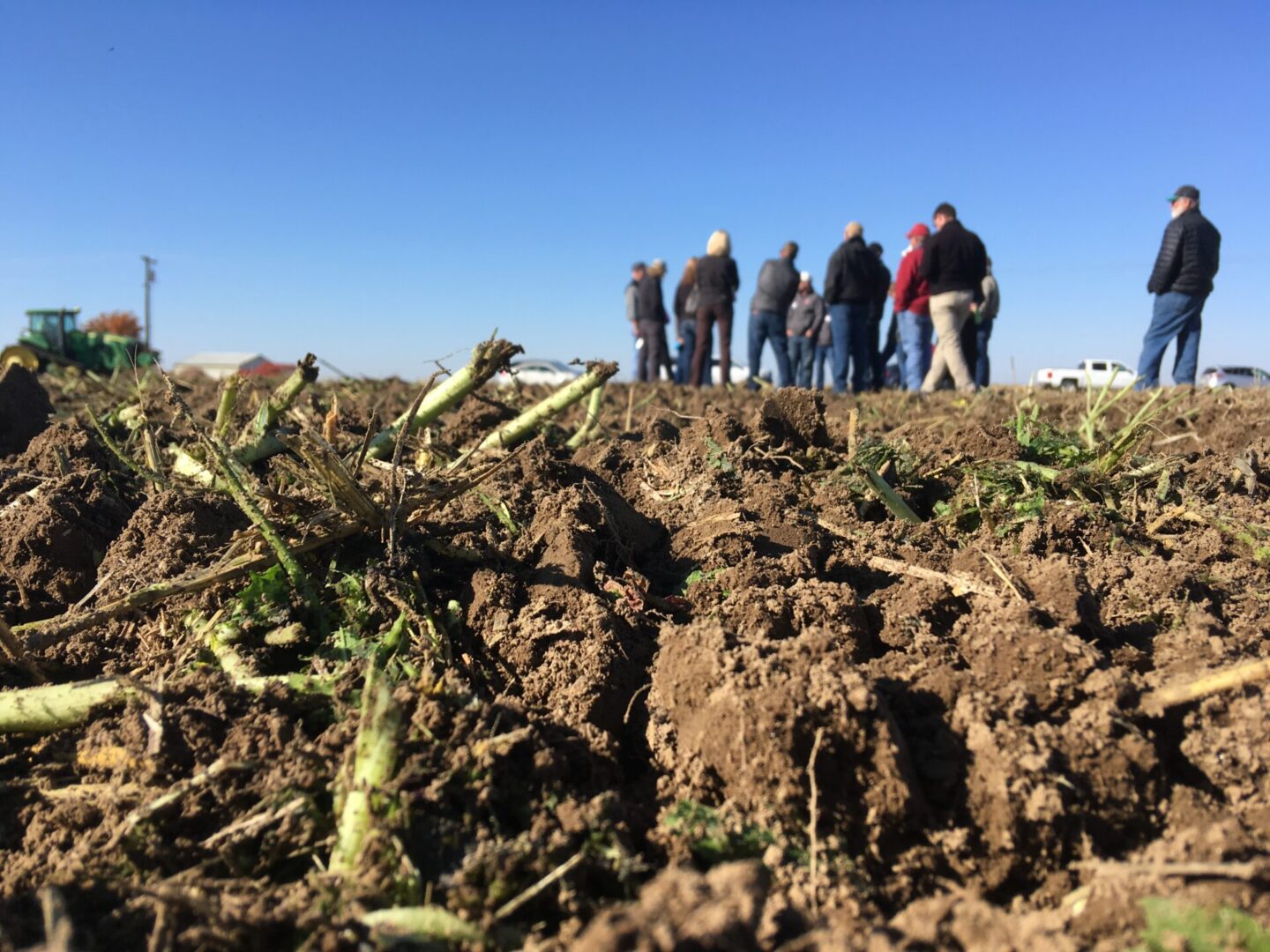 Close-up view of rough, freshly tilled soil with plant residue in the foreground. In the distance, a group of people stand together on the field under a clear blue sky, with farm equipment and vehicles visible in the background.