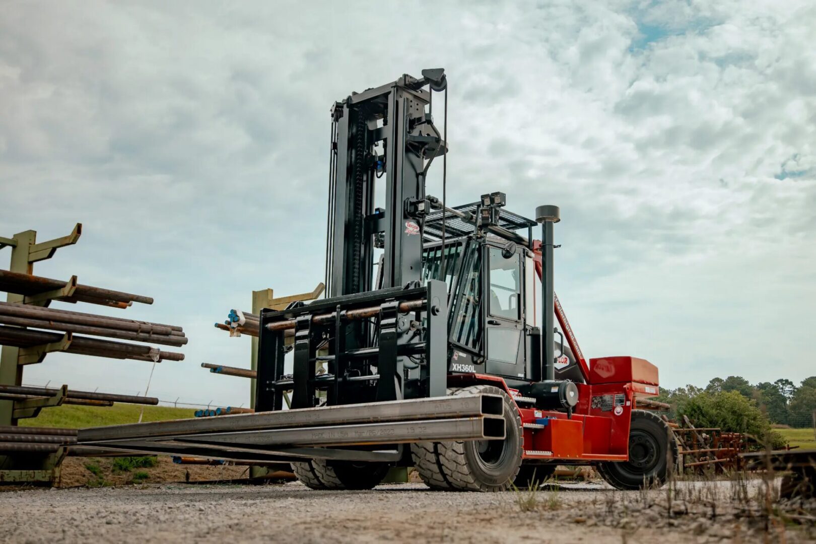 A large red-and-black industrial forklift lifts several long rectangular steel beams in an outdoor storage yard. More stacked metal beams sit on racks in the background under a partly cloudy sky.