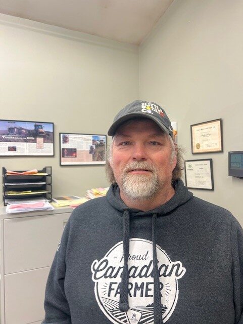 Person wearing a dark baseball cap and a dark hooded sweatshirt with the text “Proud Canadian Farmer” stands in an office. The office contains a filing cabinet, stacked paper trays, framed certificates, and agricultural photos or articles displayed on the walls. A computer monitor is mounted on the wall to the right, and the room is lit with soft indoor lighting.