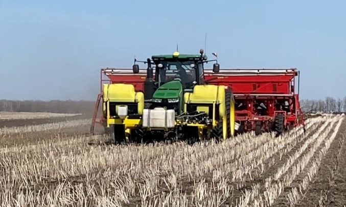 Large agricultural tractor pulling a multi-row planting machine across a harvested field. The green tractor is equipped with yellow tanks and is towing a red implement that spans several rows. The machine is traveling along evenly spaced crop rows through a field covered with short stubble under a clear blue sky, with a line of bare trees visible in the distance.