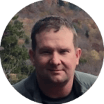 Alt text: Head-and-shoulders outdoor portrait of a person wearing a dark shirt and gray-green jacket. The person is centered in the frame and looking toward the camera. Behind the person is a wooded hillside with autumn-colored trees in shades of orange, brown, and green. The image is closely cropped around the face and upper torso, with soft natural daylight and a slightly blurred background.