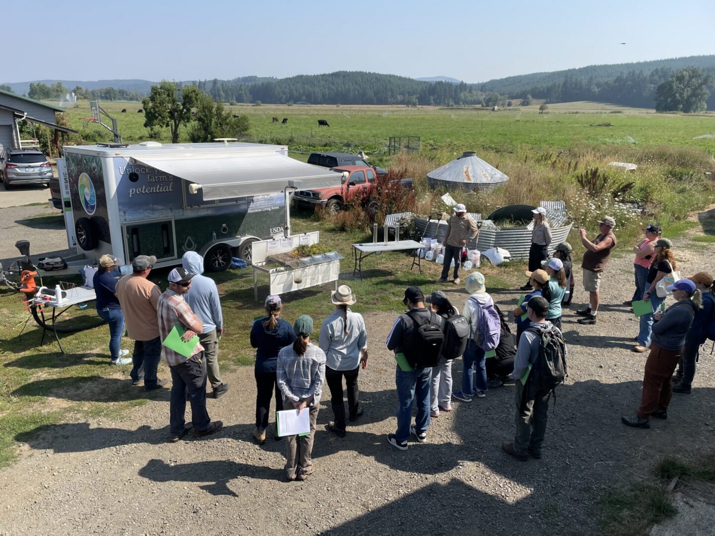 A group of approximately 25 people gathers outdoors around a USDA soil health trailer and rainfall simulation demonstration during a farm field day. Attendees stand in a semicircle listening to presenters near demonstration tables and equipment. The event takes place in a rural agricultural setting with open pasture, grazing livestock, parked vehicles, wildflower vegetation, and forested hills in the background. The scene is brightly lit by sunlight and provides an overview of hands-on soil health education taking place on a working farm.