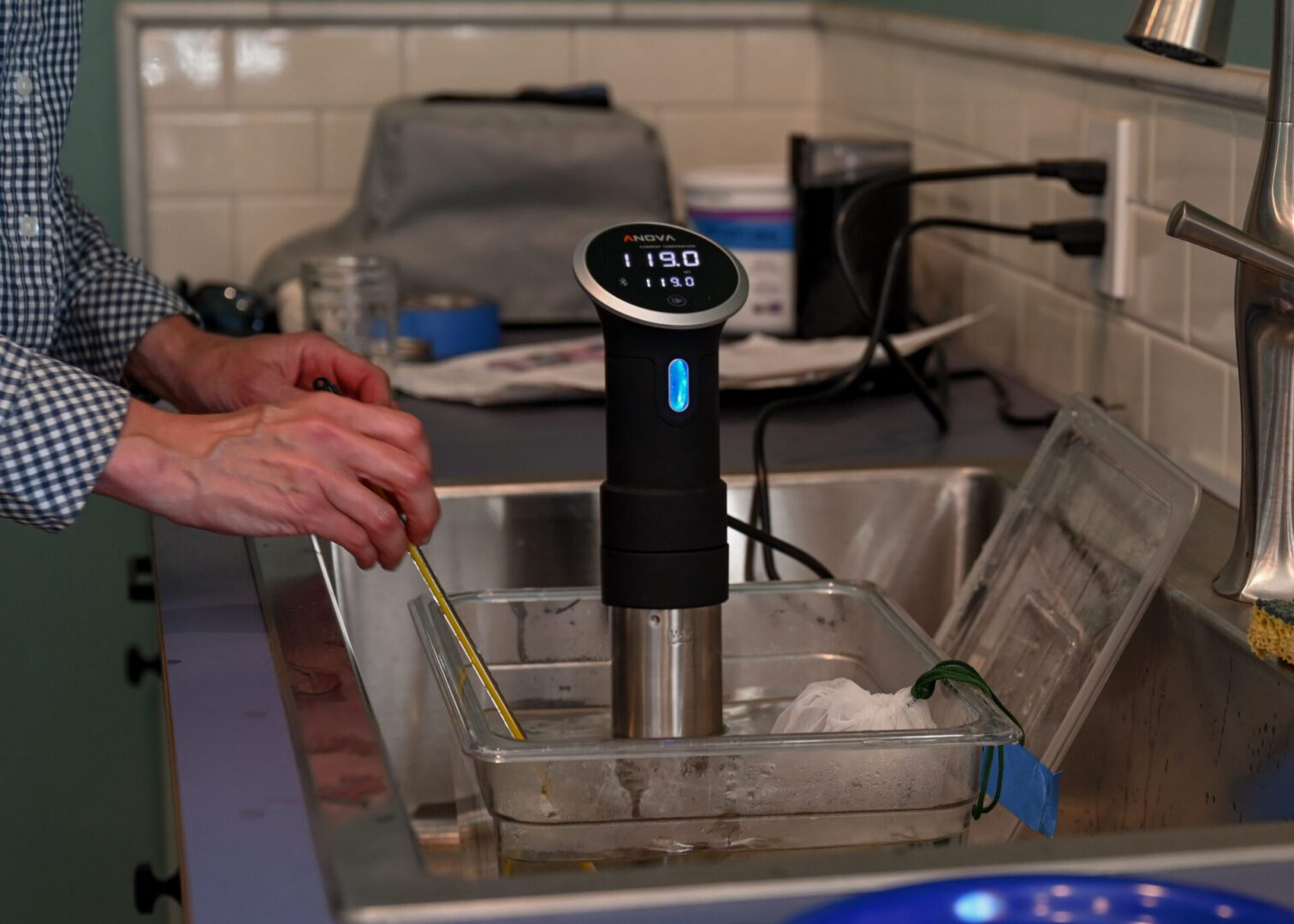 Person using a sous vide immersion circulator in a water bath set inside a kitchen sink while monitoring the temperature with a thermometer.