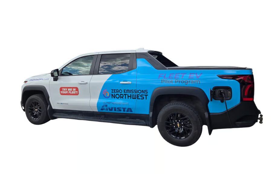 Alt text: Side view of a blue and white electric pickup truck displayed on a white background. The truck is branded with logos and promotional text for the Zero Emissions Northwest Fleet EV Pilot Program. A charging cable is plugged into the charging port on the rear passenger side. Visible text on the vehicle includes “TRY ME IN YOUR FLEET,” “ZERO EMISSIONS NORTHWEST,” “In Partnership with Avista,” and “FLEET EV Pilot Program.” The truck has black wheels, tinted windows, and a covered cargo bed, highlighting its use as a fleet-oriented electric vehicle for transportation and sustainability demonstrations.